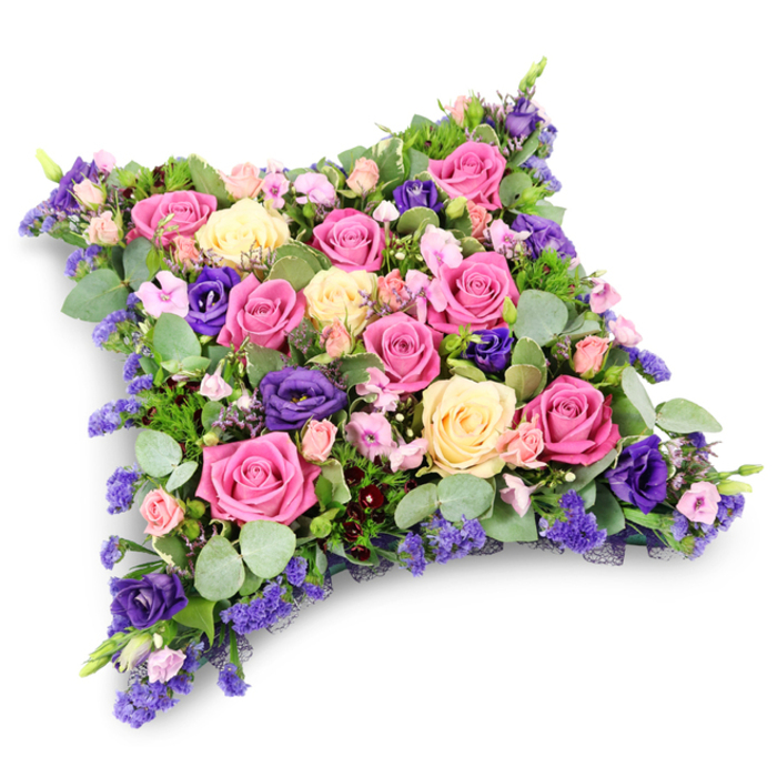 This image shows a beautifully composed funeral cushion tribute, crafted in the style of a soft pillow and set against a clean, white backdrop that allows every bloom to be clearly seen. The design forms a low, rectangular cushion with rounded corners, densely covered in a tapestry of flowers with no empty spaces, creating both visual and emotional fullness. Dominating the arrangement are large-headed roses in a thoughtful blend of rich fuchsia, gentle blush pink, and creamy pale yellow, many of them fully open so that their spiralling petals are visible down to the heart of the bloom. Interlaced between these classic roses are deep violet-purple lisianthus flowers, their ruffled, layered petals introducing a velvety texture and a more saturated depth of colour. Throughout the piece, smaller spray roses in soft pink nestle against clusters of fine, lavender-blue filler flowers, likely statice or limonium, which bring a feathery lightness and subtle, misty haze of colour to the arrangement. Around and beneath the blooms, an assortment of greenery adds structure and contrast: rounded eucalyptus leaves in a silvery-green tone soften the palette, while darker, fern-like foliage provides a more defined framework, supporting the cushion shape and adding movement along the edges. The overall colour combination of pinks, creams, and purples against lush green foliage creates a sense of gentle warmth and quiet dignity, reminiscent of a peaceful walk through nearby parks and gardens in the Tufnell Park area. The lighting is soft and even, without strong shadows, further enhancing the impression of calm. This floral pillow clearly reads as a sympathy or memorial tribute, ideal for resting beside a casket, on a memorial table, or at a graveside, conveying comfort, remembrance, and respectful farewell.