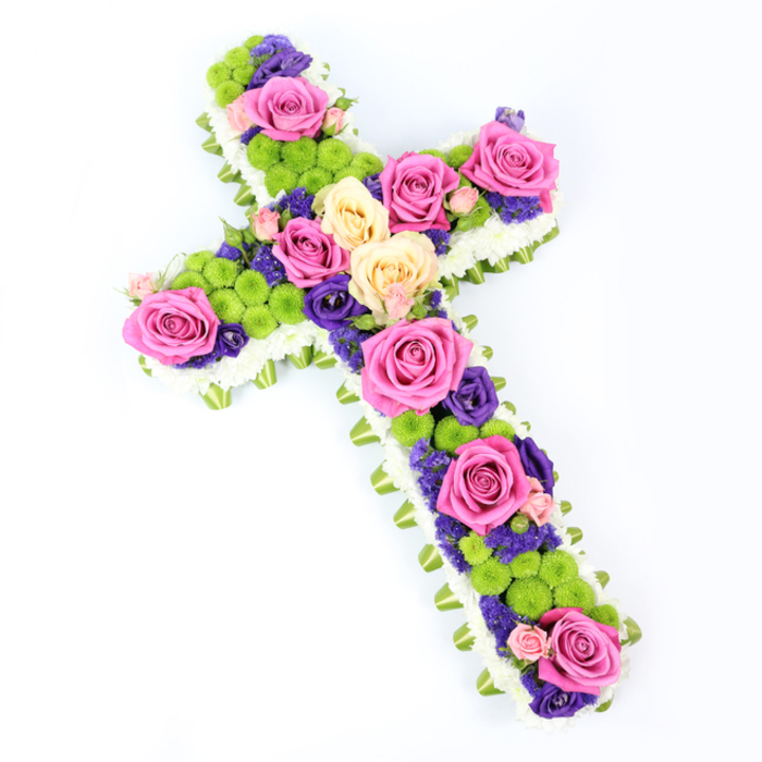 A beautifully composed floral cross fills this photograph, its full length captured against a spotless white background that highlights every blossom. The cross is positioned at a slight diagonal, almost as if it is resting on a table and viewed from above near the shoulder, with the top of the cross leaning modestly to the left and the base stretching to the right. Along the outer edge runs a continuous border of crisp white spray chrysanthemums, packed closely together to form a plush, cloud-like frame that defines the sacred shape. Just beneath this snowy edging, a subtle ring of pale green material-likely folded satin ribbon or neatly wrapped foliage-creates a fine band of soft colour, adding structure and a gentle glow to the design, similar to the way a careful florist in Tufnell Park might finish a tribute for a local church near Dartmouth Park. Inside this refined frame, the cross is filled with a lavish medley of blooms. Large, open roses in bold fuchsia pink and creamy pale yellow create strong focal points at intervals along the arms and centre, their petals layered like delicate silk. Between them, clusters of bright lime-green button chrysanthemums introduce a lively, almost playful texture, each spherical head adding depth and visual rhythm. Nestled among these larger flowers are smaller, deep purple or violet blooms-probably statice or lisianthus-which thread through the arrangement, providing rich contrast and a sense of solemn beauty appropriate for a funeral or memorial. The bright, even lighting reveals every nuance of colour and detail, from the veining in the rose petals to the tiny florets of the mums. The overall effect is of a carefully considered sympathy cross, at once dignified and vibrant, suitable for honouring a loved one from the Tufnell Park community with grace and colour.