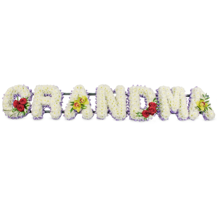 The image shows a lovingly designed funeral flower display spelling out GRANDMA in large, ornate block letters, arranged in a neat horizontal line on a clean white background. Each individual letter is constructed from a dense carpet of small, pristine white blooms, most likely chrysanthemums, their rounded petals packed closely together to create a soft, even texture with no gaps. Beneath this white surface, a delicate band of pale lavender or light purple flowers provides a subtle outline for every letter, giving just enough contrast to define the shapes while preserving a gentle, restful mood. Carefully chosen accent flowers personalise the piece. On the lower left curve of the 'G', three deep red roses with velvety petals sit among deep green leaves, offering a striking yet tasteful touch that suggests enduring love. The first 'A' on the left is decorated on its central bar with two yellow-green orchids, their lightly patterned throats drawing the eye and bringing a hint of contemporary elegance. The 'R', 'N', and 'D' remain composed of pure white blooms and the lavender trim alone, creating a calm, unembellished rhythm through the centre of the word. In the lower right section of the 'M', another cluster of three red roses mirrors the roses on the 'G', and the final 'A' repeats the first, with two more yellow-green orchids positioned carefully to complete the sequence. All of the letters are supported by a slim, dark green or black metal frame that links them at the back, ready to rest on trestles or an easel at a chapel of rest, church, or graveside in Tufnell Park or close to Tufnell Park Station. The arrangement radiates peace, affection, and gratitude, an eloquent floral tribute for a beloved grandmother.