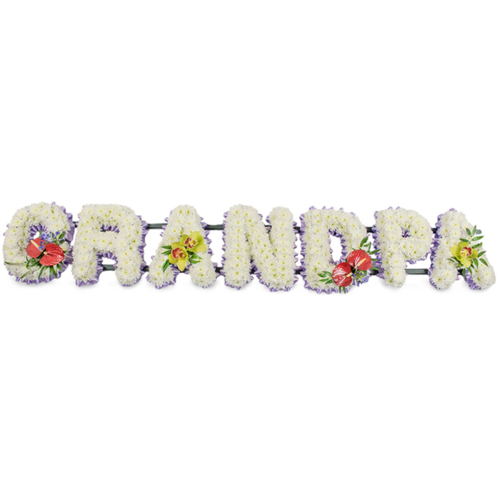 Against a crisp, uncluttered white background, a long floral arrangement spells out GRANDPA in striking, three-dimensional block capitals, each letter lovingly built from masses of pristine white chrysanthemum heads. The flowers are laid so tightly together that no gaps show, forming a velvety, almost quilted texture that reads as calm and comforting from a distance. Along the base of each sturdy letter, a soft lavender ribbon peeks through, creating a gentle band of lilac that outlines and separates the forms while introducing a quiet, respectful hint of colour. The letters rest on dark green metal or wooden rails that run unobtrusively along the back, ensuring the tribute is stable enough to be displayed beside a coffin or near the lectern during a funeral service in Tufnell Park or nearby Dartmouth Park. Carefully placed accent sprays break the white expanse with vivid touches of colour. On the G and D, bold red anthuriums emerge, their glossy, heart-shaped blooms and pronounced golden spadices nestled in rich green foliage and a few tiny purple flowers, suggesting deep love and remembrance. On the first A and the final A, clusters of yellow-green orchids create a lighter, more ethereal contrast; their elegant, star-like petals and subtle markings feel delicate yet enduring, framed by fresh leaves. This bespoke funeral flower lettering would typically be ordered by close family to honour a much-loved grandfather, perhaps placed on a hearse travelling through Tufnell Park's tree-lined streets, its careful craftsmanship and balanced design quietly expressing respect, affection, and a lifetime of shared stories.