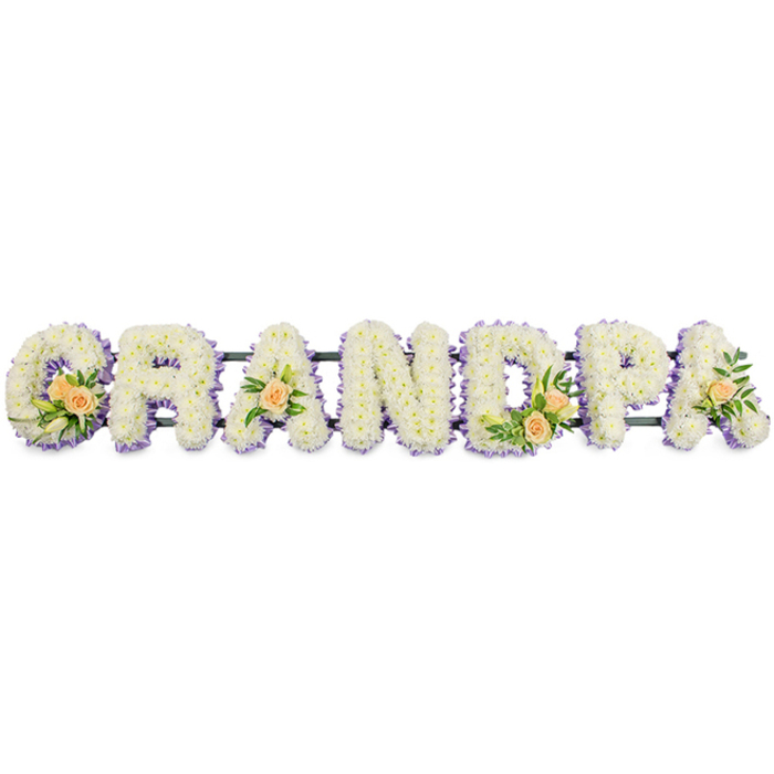 A bespoke GRANDPA funeral tribute is captured in this image, designed as a horizontal row of seven large, upright letters, each one lovingly shaped from fresh flowers with the precision of a skilled florist. The characters G, R, A, N, D, P, and A stand side by side along a slender dark support bar, creating a strong, readable word that would be easily seen at the front of a service in Tufnell Park. Every letter is generously covered with small, pristine white chrysanthemums or similar button blooms, placed so closely together that no gaps show, resulting in a smooth yet softly textured surface. Around the outside of each letter runs a consistent ribbon of pale purple flowers, a gentle lavender outline that defines the typography and lends a subtle, spiritual calm to the arrangement. These borders form a quiet frame for the white centres, allowing the word to stand out distinctly against the pure white background of the photograph. Interspersed along the tribute are clusters of peach roses and greenery that add warmth and personality: the initial G is decorated with a fuller grouping of apricot-toned roses, white buds, and rich green foliage; the first A and the final A each feature a single rose with leaves; and the D bears another small cluster, mirroring the balance of the design. These peach accents feel like intimate touches, as if each rose represents a shared memory or story. The overall composition conveys serenity and affection, a personalised floral arrangement that could be placed by family members as a final message of thanks and love to their grandfather, perhaps after a quiet walk from nearby streets and gardens in Tufnell Park.