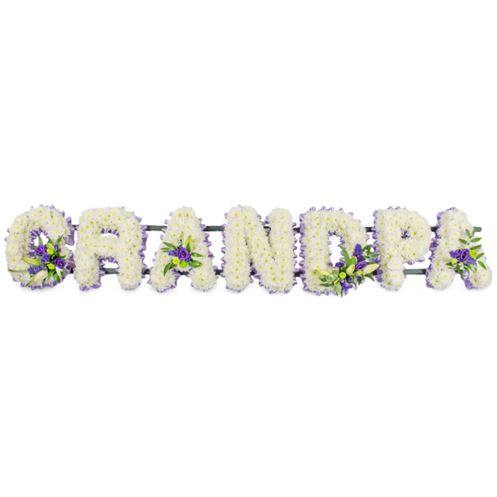 This photograph presents a personalised funeral flower tribute that spells out the word GRANDPA in bold, individual block letters, all aligned in a neat horizontal row against a pure white background. Each letter has been meticulously covered in masses of small white chrysanthemums, the petals tightly packed together so that no gaps or mechanics are visible. The surface looks pillowy and soft to the touch, evoking the idea of a gentle rest and the purity of cherished memories. Nestled carefully into the white backdrop are clusters of deep purple and lavender flowers, accented with short stems of leafy greenery. These coloured details are thoughtfully placed: a rich cluster sits in the hollow of the 'G', another nestles in the centre of the first 'A', more appear along the straight line of the 'D' and throughout the bowl of the 'P', and a final flourish adorns the last 'A'. The purple tones add quiet depth, suggesting dignity and respect, while the green foliage brings a natural, garden-like feel reminiscent of the green streets and small parks around Tufnell Park. Beneath the row of letters, a slim, dark green framework supports the design yet remains mostly concealed beneath the generous cover of blooms, ensuring the tribute appears seamless when viewed from the front, whether resting beside a coffin or displayed near the front of a service. It is easy to imagine this specific GRANDPA tribute being delivered from a local Tufnell Park florist to a chapel in nearby Holloway, serving as a focal point of remembrance. The arrangement combines classic funeral whites with soothing purples to express love, gratitude, and the enduring presence of a much-missed grandfather.