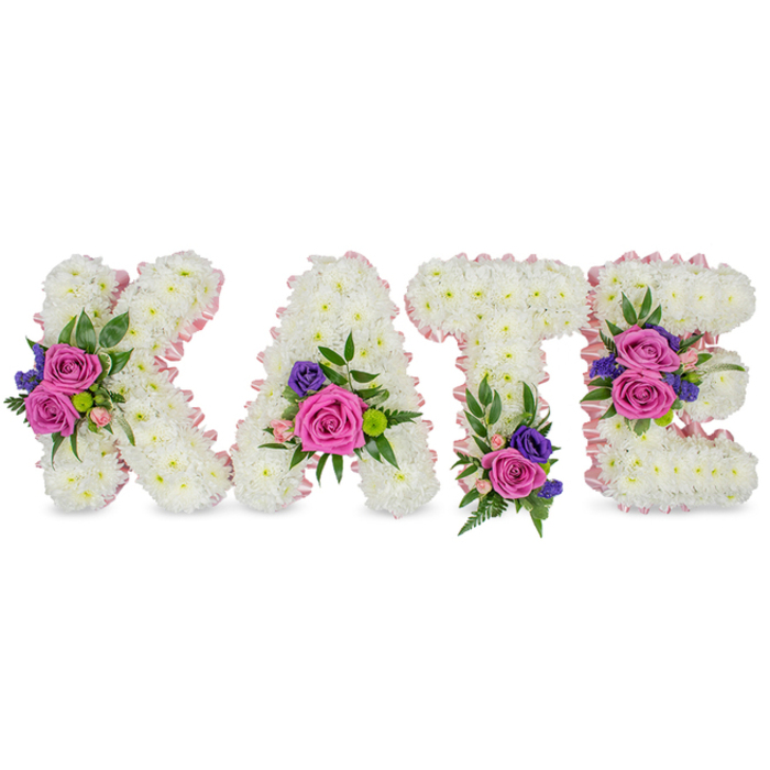 A lovingly made standing tribute spells the name KATE in four separate, upright letters, each letter built from a thick, carpet-like layer of pure white chrysanthemums that read as satin-soft to the eye. Pale pink satin ribbon traces the contours of each character, providing a subtle halo effect that warms the stark white surface and defines the letterforms with gentle elegance. Accents of deep fuchsia-pink roses and smaller pastel pink spray roses are arranged in harmonious clusters alongside cooler purple-blue accent flowers-probably statice or lisianthus-so that bright, velvety petals sit against cool-toned fillers for dramatic but soothing contrast. The clusters are nest-led in varied greenery: fern fronds add a feathery, tactile element while eucalyptus leaves contribute smooth, elliptical shapes and a subdued aromatic note.

Design details read like a careful map of memory: a bold cluster adorns the diagonal of the K, the A bears flowers near its lower right curve, the T is weighted with a bouquet at its base, and the E shows two distinct groupings on its middle and lower bars. The palette of crisp white, fuchsia, soft pink, and deep purple reads tender and respectful-perfect for a funeral service, graveside tribute, or celebration of life. The lighting in the image is soft and neutral, revealing the chrysanthemums' texture and the roses' velvety sheen without harsh shadows. This bespoke floral letter arrangement would sit comfortably at a chapel on Tufnell Park Road or a quiet service near Dartmouth Park, offering a personalized and heartfelt way to honour someone named Kate.