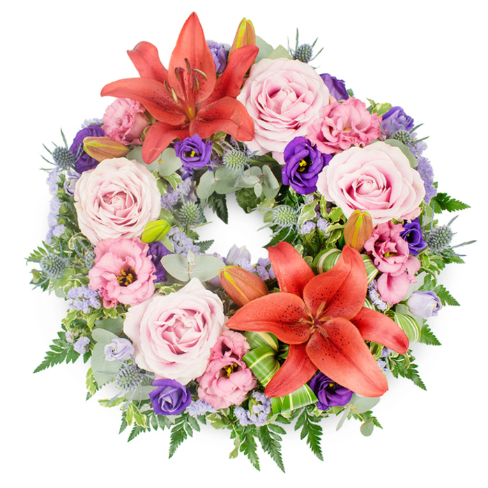 Against a plain white backdrop, a richly detailed sympathy wreath lies perfectly circular, inviting the eye to travel slowly around its ring of colour and texture. The arrangement is dense and deliberate, as though every flower has been carefully placed by an experienced Tufnell Park florist used to guiding families through difficult goodbyes. Soft pink and blush roses appear throughout the circle, many fully open so that their spiralling centres are clearly visible, giving a sense of fullness and grace. Beside them, eustoma blooms in both pale pink-lilac and intense, royal purple add variation, their gently ruffled petals providing a soft contrast to the sleek rose heads. Two bold coral-orange Asiatic lilies, one positioned slightly off-centre near the top and another echoing it at the lower edge, form strong focal points; their wide petals and central stamens catch the light and add a subtle vibrancy to the otherwise muted palette. Tiny lavender-blue filler flowers, likely statice or limonium, weave in and out between larger blooms, like a fine veil overlaying the design. The base of the wreath is built from an intricate mix of greenery: round, silvery-green eucalyptus leaves, spiky blue eryngium thistles, feathery ferns, and darker, more compact foliage that anchors the ring visually. A few unopened lily buds peek from the greenery, suggesting blooms yet to open, like memories that remain vivid. Near the bottom lily, bright green, folded leaves, shaped perhaps from aspidistra or Ti leaves, are arranged in crisp loops that highlight the artistry of the florist. This wreath, suitable for a funeral service in Tufnell Park or a memorial gathering in nearby local venues, carries an atmosphere of calm remembrance and respectful farewell.
