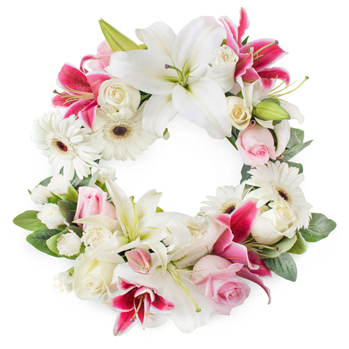 An open circular funeral wreath is presented on a crisp white background, its generous ring of flowers forming a serene and balanced tribute. The palette is soft yet expressive, built around shades of white, blush pink, and rich magenta. At first glance, the eye is drawn to the large white lilies spaced throughout the circle, their broad, silky petals opened wide, exposing delicate green pistils and brushed-gold stamens that lend a sense of quiet radiance. Nestled between them are deep pink and fuchsia lilies, their star-shaped blooms and subtle speckling bringing warmth and contrast, like a gentle echo of colour you might see on a calm evening walk through the residential streets of Tufnell Park. White gerbera daisies with dark chocolate or reddish-brown centres punctuate the wreath, their tidy, layered petals adding a playful but still respectful texture. Soft, light pink roses mingle with creamy white roses, some still in bud and others blooming fully, creating a tapestry of petals that suggests love, devotion, and remembered tenderness. Smaller white filler flowers-possibly miniature carnations or clusters of spray roses-are threaded throughout, closing any gaps and giving the wreath a full, plush appearance. The blooms sit on a bed of lush, varied greenery, with glossy leaves and subtle buds providing depth and framing the circular form. The overall impression is of a carefully composed, heartfelt tribute, perfect for a funeral, wake, or memorial service in or around Tufnell Park. Crafted with the care of experienced local florists, it conveys purity, respect, and enduring affection for the person being honoured.