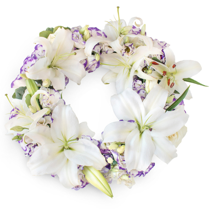 A full, meticulously crafted sympathy wreath is shown against a clean white backdrop, the circular shape immediately communicating continuity and the enduring nature of love and memory. The wreath is densely woven with large white Oriental lilies, whose star-shaped blooms stretch outward with an almost weightless elegance. Each lily displays six broad, velvety petals, slightly reflexed at the tips, creating gentle shadows and highlights under the soft, even lighting. Their centres glow subtly with a greenish-yellow hue, from which upright pistils and delicate stamens emerge, some carrying golden or rust-coloured pollen that offers a warm contrast within the otherwise cool palette. Interlaced throughout the lilies are clusters of Lisianthus, their dainty, multilayered petals frilling at the edges and tipped in shades of lavender and violet, echoing the soft purple tones often associated with remembrance and dignity. A few tight buds and smaller creamy blooms nestle close to the open flowers, adding to the sense of depth, like quiet thoughts tucked in among spoken words. Slender green leaves and eucalyptus-style foliage wind discreetly beneath the blossoms, offering structure and a calming, botanical freshness. The composition is balanced and symmetrical, yet not rigid, evoking the gentle, natural grace of a thoughtfully tended garden. This wreath feels particularly suited to a funeral or wake in Tufnell Park, perhaps accompanying a service near the station or one of the tree-lined side streets that give the area its village-like calm. Crafted with the kind of care local florists bring to every farewell, it provides a refined, heartfelt tribute for families seeking a white and purple wreath that reflects both purity and soft, enduring grief.