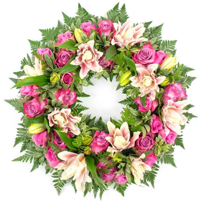This image presents a meticulously crafted circular funeral wreath, isolated against a bright white background so that every bloom and leaf is clearly defined. The wreath is composed of a continuous ring of flowers, dominated by roses in shades of deep magenta, saturated fuchsia, and more delicate, powdery pink. The roses vary in openness: some form tight, spiralled buds suggestive of new beginnings, while others are fully open, displaying many layered petals that curve and fold inwards, creating a sense of depth and gentle movement. Interwoven through the roses are large double lilies whose broad, velvety petals are coloured in creamy ivory, blush pink, and light peach, with occasional deeper pink accents at the tips or along the veins. Several lime-green lily buds punctuate the design, their pointed forms providing contrast to the rounded rose heads. Surrounding and underpinning the blooms is an abundance of foliage, including finely textured fern fronds that radiate softly from the outer edge and glossy, dark green leaves that offer structure and a rich background. Some leaves catch the light with a subtle sheen, while others are matte, adding further textural interest. The composition is full and symmetrical, with no gaps, giving a feeling of completeness and continuity that is often sought for memorial tributes. This wreath would be perfectly at home at a funeral chapel in Tufnell Park, carried along Junction Road, or placed beside a memorial in nearby Waterlow Park, where its pink and cream tones would stand as a tender symbol of love, respect, and cherished memories for the person being honoured.