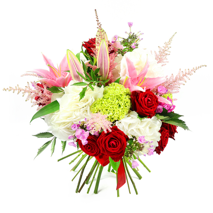 A richly detailed hand-tied bouquet is centred against a crisp white background, the entire composition designed to feel both opulent and welcoming, like a gift arriving at a Tufnell Park doorstep on a special afternoon. Several deep red roses dominate the middle of the design, their velvety petals folded in tight, romantic whorls that immediately signal love and admiration. Clustered nearby, generous pink lilies rise above the other flowers, with some wide open to display their stamens and pale inner throats, while others remain as elegant, tapered buds edged in a soft green tinge, hinting at fragrance yet to be released. Around these stars of the show, ample heads of ruffled white blooms-reminiscent of hydrangeas or full carnations-form a creamy, cushion-like base, adding volume and a sense of pure, gentle light. A vivid, cushion-shaped cluster of bright lime green dianthus sits slightly off-centre, its fine, frilly texture creating a striking contrast to the smooth rose petals and giving the arrangement a fresh, contemporary edge that feels at home in the creative corners of Tufnell Park. Wisps of pale pink astilbe stretch outward from among the larger blossoms, their feathery plumes lending movement and a soft, romantic haze, as if a light mist has settled over a summer garden. Tucked in between are small fuchsia and lavender blossoms, subtle yet effective in deepening the colour palette and guiding the eye from one bloom to the next. Dark green foliage peeks through at various points, glossy leaves framing and grounding the brighter tones. At the base, the green stems are gathered tightly and bound with a sleek red satin ribbon, completing the bouquet's polished, gift-ready look. This arrangement perfectly suits anniversaries, milestone birthdays, or simply brightening a living room overlooking Tufnell Park station, while reflecting the care and craftsmanship of local florists.