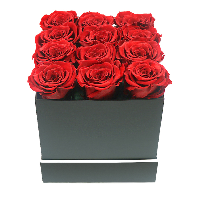 The photograph captures a meticulously arranged square box of red roses, styled in a way that feels at once modern and timeless, much like the evolving streets around Tufnell Park. At the centre of the image sits a compact, matte black square box, its surfaces perfectly smooth and its edges sharply defined, giving it the clean-lined look of a contemporary gift. Rising from the open top are twelve sumptuous red roses, aligned in a precise 3x4 pattern, each bloom pressed closely to the next so there are no gaps between them. The roses are in full, generous flower, with layered, velvety petals that spiral outwards in deep crimson tones, from near-black shadows nestled in the centre to brighter ruby highlights at the outer edges. The blooms extend slightly above the rim of the box, creating a plush, cushion-like effect that suggests abundance and care. A slender white band encircles the lower section of the box, introducing a crisp, graphic line that contrasts beautifully with the rich black surface and intensifies the visual impact of the red flowers. The arrangement is photographed against a spotless white background, with gentle, even lighting that minimizes harsh reflections and allows the texture of each petal to be clearly defined. Subtle shadows beneath the petals and along the base of the box add depth and dimension, so the arrangement appears almost three-dimensional on the page or screen. There are no additional decorations or foliage, reinforcing a minimalist, luxury aesthetic perfect for same-day romantic deliveries around Tufnell Park, Archway, or Gospel Oak. This composition conveys enduring affection, refined taste, and a preference for thoughtful, long-lasting floral gestures.