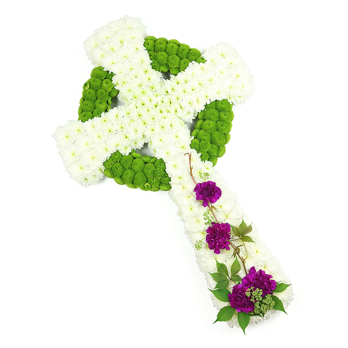 A meticulously designed Celtic cross floral tribute dominates this clean white studio image, the arrangement turned slightly on the diagonal from top left to bottom right to emphasise its full shape. The cross is almost entirely surfaced with petite, snow-white chrysanthemums, their tight, rounded heads pressed so closely together that they form a continuous, velvety carpet of petals along both the vertical and horizontal arms. Set into this white canvas is the traditional Celtic ring and patterning, crafted from striking lime green button chrysanthemums that create a bold, circular halo at the centre and subtle knot-like shapes that echo down the arms. These green flowers sit just a touch higher than the white, adding layered texture and visual depth, so the design appears almost carved in flowers rather than stone. On the lower portion of the upright arm, three distinct clusters of deep purple carnations introduce a poignant burst of colour. Each cluster is slightly different in size, arranged so the blooms seem to tumble softly down the cross, their many ruffled petals rich and velvety. Fine strands of dark green foliage, reminiscent of ivy or delicate woodland greenery, weave among the carnations and stretch lightly over the white chrysanthemum surface, suggesting a sense of nature gently reclaiming a sacred symbol. The lighting is bright and even, highlighting every nuance of petal and leaf. This floral cross feels especially suited to a memorial steeped in tradition, perhaps chosen by a family from the Tufnell Park or Archway area who wish to honour Irish or Scottish roots, or simply to express steadfast faith and peace through a beautifully crafted sympathy tribute.