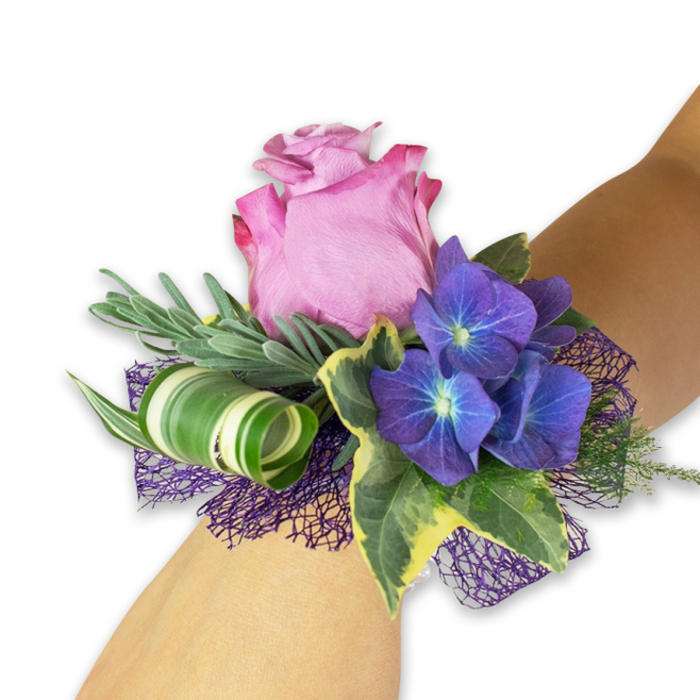 Set against a crisp white field and resting on a pale wrist, this wristlet reads as a study in texture and colour harmony, perfectly scaled for special-occasion wear. A single, partially open pink rose commands the centre with lush, velvety petals that graduate from a soft blush at the heart to a more saturated fuchsia-magenta at the ruffled tips-an elegant, romantic anchor. Flanking the rose are dense clusters of deep indigo-purple hydrangea; each small floret is punctuated by a faint blue-white centre, creating an almost luminescent effect that adds dimensional depth and tactile contrast. Long, narrow silvery foliage-likely eucalyptus or lavender-fans out behind the blooms, introducing a cool, smoky green and a whisper of fragrance. Variegated ivy leaves in deep green with creamy yellow margins provide classic structure and glossy sheen, and a broad, striped leaf curled into a deliberate spiral gives the composition a contemporary architectural note. Below and around the stems, a sheet of dark purple, open-weave mesh peeks through, lending a soft, netted cushion that amplifies the hydrangea's tones while keeping the arrangement light and wearable. The overall impression is refined and celebratory; I picture this corsage arriving at a prom near Junction Road, adorning a bridesmaid at a small Tufnell Park wedding, or accompanying a graduate heading to a nearby ceremony. The craftsmanship is clear in the balance of soft blooms, sculptural foliage, and textured backing, designed for comfort and visual impact.
