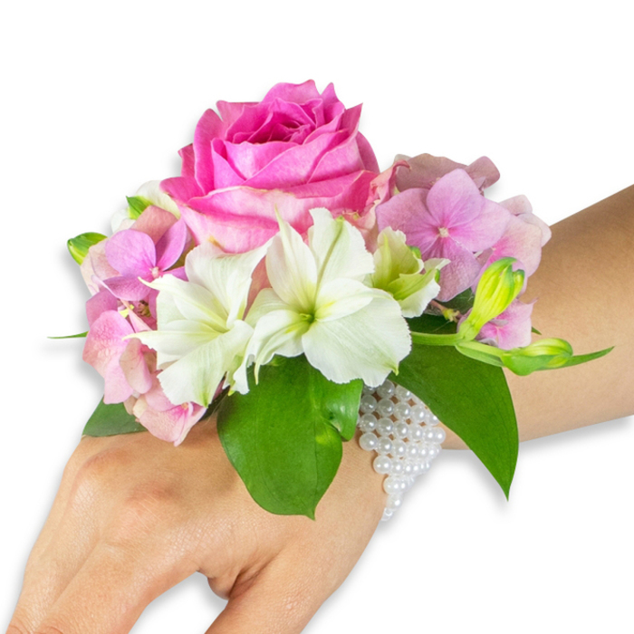 Presented on a light-to-medium skin-toned left wrist and forearm against a stark white backdrop, this corsage is a study in refined proportion and texture that would sit comfortably at a Tufnell Park wedding or a celebratory evening on Tufnell Park Road. The arrangement centres on a large, fully-bloomed hot fuchsia-pink rose whose dense, overlapping petals form a rich, tactile heart. Surrounding that focal rose, delicate clusters of soft, blush-pink hydrangea florets create a voluminous, cloud-like base; their small, rounded petals add a gentle, luminous mass that contrasts with the rose's structural depth. Interwoven are several white alstroemeria (Peruvian lilies), each with four to six slender petals and faint light-green striping near the throat, lending crispness and an elegant, slightly botanical silhouette. On the arrangement's right flank a sprig of light green alstroemeria buds and a few almost-closed pink buds suggest movement and the quiet patience of nature. The composition rests upon several broad, glossy emerald leaves that anchor the colour story and provide a rich backdrop to the pastel blooms. Securing the piece is a delicate, multi-strand bracelet of small, pearlescent white beads; the pearls wrap the wrist in a refined net and trail delicately toward the hand, catching studio light with a soft, satiny glimmer. The overall effect is both youthful and classic, an expertly constructed wrist corsage suited to proms, bridal parties, and intimate celebrations around Junction Road and nearby neighbourhood venues.