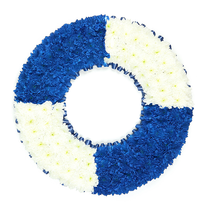 This image shows a beautifully composed funeral wreath crafted in the unmistakable shape of a lifebuoy, set against a crisp white background that keeps the focus firmly on the flowers. The circular tribute is meticulously packed with small, uniform blooms-most likely chrysanthemums or similar button flowers-arranged so closely that they form a smooth, padded surface, almost like fabric. The ring is visually divided into four quarters, alternating between deep royal blue and pure white segments. Starting from the upper left, a bold square of blue blooms curves around the form, followed by an equally dense section of white at the top right. The pattern repeats at the bottom, with another sweep of intense blue and then fresh white, creating a rhythmic maritime colour blocking reminiscent of traditional lifebuoys found by the water. Each petal is carefully placed, and the high level of craftsmanship shows in the perfectly even outer and inner circles. Along the inner rim, a dark blue ribbon has been folded and ruffled to create a delicate border of soft pleats, echoing the main blue quadrants and adding an extra touch of refinement. The central hole remains clearly open, emphasising the lifebuoy symbolism and creating a calm, contemplative space within the design. As florists serving the Tufnell Park community, we often see families choose such a piece to honour loved ones connected to the navy, coast guard, or those remembered as a 'lifeline' to others. The stark white background, much like the pale walls of many homes near Tufnell Park station, heightens the visual impact of the vivid blue and serene white flowers, making this a poignant and memorable tribute for local funeral services and chapel gatherings.