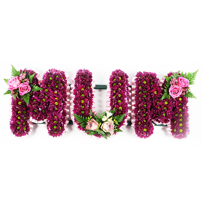 The image shows a lovingly designed floral tribute where the word MUM is created in generous, capital letters, standing out clearly against a crisp white backdrop. Each letter is formed on a solid base and then completely cloaked in hundreds of compact spray chrysanthemums in rich tones of magenta, fuchsia-purple, and deep pink. The flowers are so closely arranged that no gaps in the structure beneath are visible, giving a plush, cushion-like surface that invites the eye to trace every curve of the typography. The green centres of the chrysanthemums appear as tiny, bright dots amid the darker petals, introducing a natural contrast that keeps the design fresh and uplifting. The sides of each letter are edged with a narrow, ruffled ribbon in the softest blush pink, carefully pinned to follow the contours of the Ms and U, adding a gentle, almost lace-like frame. On the upper left of the first M, two open pink roses and several rosebuds nestle among flowing fronds of fern and glossy greenery, creating a focal accent that feels both elegant and heartfelt. At the base of the central U, a trailing crescent of paler blush roses, creamy blooms reminiscent of freesia, and fern leaves rests like a small garland at the feet of the letter. The second M echoes the first with a matching cluster of bright pink roses and foliage on its upper right edge, bringing the composition neatly into balance. It is the kind of bespoke sympathy tribute a family in Tufnell Park might place at a graveside or crematorium, a tender floral expression of love and remembrance for a cherished mother.