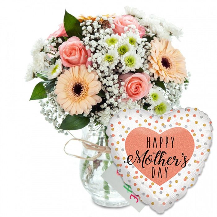 A warm and celebratory Mother's Day composition is photographed against a clean white background, giving the feeling of standing in a bright, airy Tufnell Park living room just after a florist's delivery has arrived. To the left of the frame, a generous mixed bouquet is arranged in a clear, cylindrical glass vase with gentle curves and a touch of rustic charm from the light brown twine tied snugly around its neck. The blooms are a soothing symphony of peaches, pinks and whites: several peach-pink roses are captured at different stages of opening, their petal edges softly curled and velvety, while two or three peach-orange gerbera daisies provide bold focal points with their large, rounded petals and deep brown centres. Between them, clusters of delicate white gypsophila create misty layers, and small daisy-like white chrysanthemums with hint-of-green centres add a crisp, fresh texture. Rich green foliage fans out at the back and sides, shaping the bouquet into a neat yet abundant dome that looks carefully crafted rather than overly formal, like something prepared by a local Tufnell Park florist who knows the neighbourhood's relaxed style. Partially hidden at the back of the arrangement is a small white greeting card, its coloured writing slightly blurred, suggesting a personal Happy Mother's Day note from a son or daughter. To the right of the vase sits a large heart-shaped foil balloon, slightly leaning forward. The balloon is bright white, sprinkled with shiny gold and silver dots reminiscent of confetti drifting over a celebration, and in the centre a broad peach heart cradles the black inscription HAPPY Mother's DAY in a mix of bold capitals and elegant script. The combination of tender flowers, thoughtful card and playful balloon captures the essence of a lovingly planned Mother's Day surprise, ready to brighten homes all across Tufnell Park and nearby Gospel Oak.