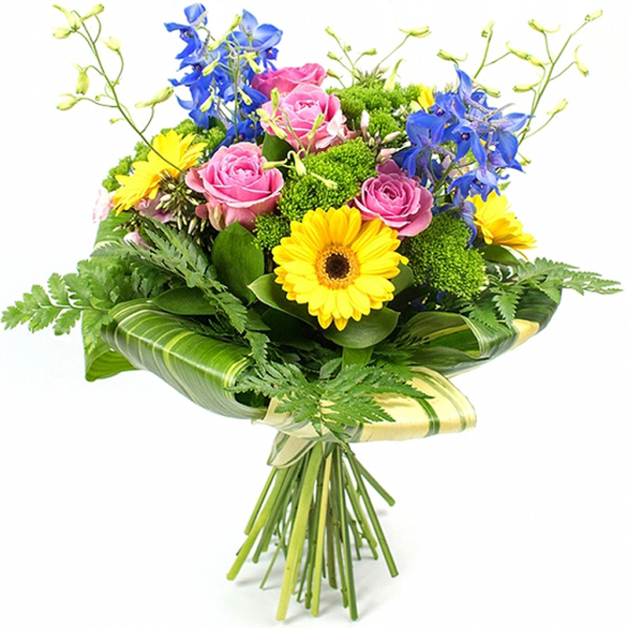 Against a crisp white background, a professionally arranged, hand-tied bouquet bursts with saturated colour and layered texture, evoking the feel of a freshly crafted design from a Tufnell Park florist's workbench. Several large pink roses take centre stage, their petals opening in gentle, layered curls that speak of tenderness and thoughtful affection. Around them, bright yellow gerbera daisies with dark brown centres create a joyful ring of daisy-like faces, adding a sunny brightness reminiscent of a clear morning over Tufnell Park Fields. Shooting upward from the heart of the bouquet, multiple stems of deep blue delphiniums introduce vertical rhythm and a cool, airy contrast, their many small florets forming a graceful column that draws the eye. Surrounding these star blooms is a rich tapestry of greenery: delicate fern fronds soften the edges and spill slightly outward, while broad, dark green leaves with a lacquer-like sheen create a strong frame that makes the colours pop even more. Between the larger elements, compact tufts of bright green filler flowers and buds add texture and volume, like cushions of fresh foliage tucked carefully by an experienced hand. Around the base of the flowers, wide tropical-style leaves, some subtly striped in lighter green, are wrapped in smooth curves to form a sculpted collar, giving the bouquet a contemporary finish often requested for celebrations and thank-you gifts around nearby Gospel Oak and Kentish Town. The natural stems extend beneath this collar, neatly gathered and bound with a pale wrap and simple tie, making the bouquet appear ready to be lifted from the image and delivered to a doorstep in Tufnell Park as a symbol of joy, appreciation, and everyday beauty.