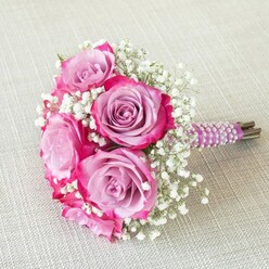 This enchanting bouquet, handcrafted by an experienced Tufnell Park florist, intertwines artistry with romantic spirit, making it a beloved choice for weddings and meaningful moments celebrated within the vibrant local community. At its core lies a harmonious blend of pink and soft lavender roses, their petals unfolding in a mesmerizing gradient from gentle mauve centers to intense fuchsia edges, drawing in the eye much like the blossoms one might see flourishing in neighbourhood gardens on a spring day. Wisps of white baby's breath thread delicately through the arrangement, lending an ethereal, airy texture that lightens the bouquet's overall form and offers a gentle counterpoint to the lush, velvety rose blooms. Everything is unified by a slender, deep purple satin ribbon, expertly wound and adorned with lines of iridescent pearls that spiral upward, adding a touch of refined opulence and tactile interest. The ribbon's sheen picks up the subtle morning light, each pearl glimmering softly, while the natural, neatly trimmed stem ends lend authenticity and a connection to the freshness of locally sourced flowers. Resting on a pale, linen-like backdrop reminiscent of the contemporary homes around Tufnell Park Road, the bouquet stands out as a radiant centrepiece, ready for a ceremony or heartfelt celebration. The arrangement's rounded, compact shape, attention to floral detail, and thoughtful finishing touches embody the dedication and creativity found in North London flower design, offering beauty and inspiration for any romantic occasion in the Tufnell Park area.