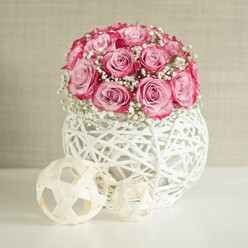 An intimate floral vignette captures a domed bouquet of pink roses and white baby's breath, styled in a way that feels perfectly at home in a Tufnell Park townhouse living room. At the centre of the image, about fifteen roses form a tight, rounded cluster, their petals unfurling in layers of colour that shift from pale, shell-like pink at the heart to a richer, fuchsia-toned rim, as if each flower holds a blush of evening light. Threads of delicate gypsophila weave between the blooms, their tiny star-shaped white flowers creating a soft mist that lightens the arrangement and adds a sense of purity and celebration, ideal for birthdays or quiet romantic gestures delivered locally. The bouquet is nestled into a distinctive spherical vase, crafted from white-painted, woven natural fibres that cross and spiral to form an open, airy lattice. Light slips through these gaps and pools gently on the smooth white tabletop below, adding to the composition's brightness. To the left sit two decorative spheres in a natural beige tone, formed from interlocked wooden strips, their geometric lines echoing the woven vase while introducing a subtle, earthy contrast. Behind, a warm beige or cream wall with a lightly textured surface serves as a calm, neutral backdrop, much like the softly painted walls in many period homes around Fortess Road and Dartmouth Park. The light in the scene is soft and diffused, revealing every curve of petal and thread of fibre without harsh shadows. The overall feeling is one of thoughtful elegance and quiet joy, like a bouquet chosen to say I'm thinking of you from a trusted local florist, then placed with care on a favourite shelf or hallway console.