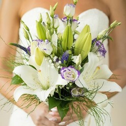 This bridal bouquet, thoughtfully arranged in the heart of Tufnell Park, presents a blend of refined artistry and romantic impulse. Central to the composition are elegant white lilies, their softly curved petals and subtle fragrance evoking the classical parks and garden squares of the area. The lilies are tenderly embraced by white lisianthus blooms, each one displaying ruffled edges tinted with gentle lavender, lending a dreamy, whimsical quality. Deep purple accents of veronica rise gracefully amid the delicate spray of baby's breath, creating a lively interplay of color and texture that is both contemporary and timeless. Surrounding the florals, wispy asparagus fern and the glossy depth of broad green leaves capture the freshness of a local spring morning, while unopened buds whisper of milestones yet to be shared. The bride's hands, carefully cradling the bouquet against the backdrop of her white gown, reflect the personal touch and care found at Tufnell Park's local weddings. Each stem has been chosen and placed with intention, forming a bouquet that tells a story of understated sophistication and optimistic new beginnings. This arrangement, perfect for couples across Tufnell Park and nearby Holloway, harmonizes tradition with modern romance-capturing the spirit of a joyful wedding day in every detail.