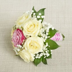 Imagine a bridal or bridesmaid bouquet that quietly pays homage to the gentle mornings of Tufnell Park, its design flowing with poetic sensibility and refined grace. Creamy roses, each with petals woven like silk, form the heart of this arrangement, their softness illuminated by the warm, natural light filtering through Victorian bay windows common in the area. Amidst these ivory blooms, one striking magenta rose is cradled just off-center-a secret jewel that draws the gaze and brings a burst of joyful color, reminiscent of the vibrant weekend blooms at the Tufnell Park Farmers' Market. Wispy sprays of white baby's breath twine in and out, evoking the delicate early mist that sometimes lingers in local gardens. Alongside the florals, broad, deep green leaves offer grounding structure and a touch of woodland freshness. Each stem is elegantly wrapped in a radiant pink satin ribbon, its smooth sheen tightly finished and entwined with a coil of luminous pearls, adding both glamour and tactile interest. The composition rests serenely on light, textured linen, amplifying the bouquet's airy beauty while underscoring its purpose-as a symbol of new beginnings and cherished moments, perfect for weddings or elegant gatherings in the heart of North London. This bouquet embodies the artistry and deep floral knowledge beloved by the Tufnell Park community, making it ideal for those seeking timeless romance.