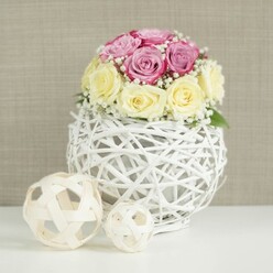 An elegant floral centerpiece sits calmly on a pristine white surface, as if ready to be placed on a wedding table in a Tufnell Park café or a stylish home near Dartmouth Park. The focal point is a compact, rounded bouquet of roses in two dreamy tones: cool lavender-pink blooms clustered beside soft, buttery yellow roses that fade into ivory at their centres. Each rose is meticulously positioned so that their ruffled petals touch and overlap, creating a lush, almost cushion-like dome of colour. Nestled between the larger blooms are dainty sprigs of white baby's breath, their tiny star-shaped blossoms forming a delicate halo that lightens the composition and adds a cloud-soft texture. Supporting the flowers is an eye-catching white spherical vase, crafted from slender branches or wicker that have been carefully interlaced and painted a clean, bright white. The open-weave structure curves into a globe, leaving gaps that hint at the mechanics inside, adding a handcrafted, slightly rustic charm while still feeling contemporary. To the left of the main vase rest two additional decorative spheres, this time in natural, pale wood tones, echoing the woven pattern and creating a harmonious trio of textures. The entire scene is framed by a neutral, beige-taupe background with a subtle fabric-like texture, similar to a refined wallpaper you might see in a renovated Tufnell Park period home. Gentle, even lighting washes across the arrangement, picking out the smoothness of each petal and the tactile lines of the wicker. The mood is serene and romantic, ideal for wedding receptions, engagement dinners, or editorial features about sophisticated table décor and same-day floral styling in North London.