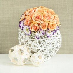 On a smooth, white tabletop that could easily belong in a townhouse just off Tufnell Park Station, a meticulously crafted floral centerpiece radiates calm elegance. A rounded dome of roses in soft peach, apricot, and salmon tones forms the core of the design, each bloom open and plush, their petals layered in generous spirals that invite the eye to linger. The roses fit together so closely that they create a seamless globe of colour, suggesting warmth, tenderness, and that gentle sense of occasion you might want for a registry office celebration or a family gathering near Holloway Road. Circling the base of this rose canopy is a collar of lisianthus flowers, their white petals delicately edged in rich violet and deep purple. These frilled blooms introduce a striking contrast, the cool tones setting off the warmth above while adding a sense of fine detail, almost like hand-painted china. Dotted between them, tiny clouds of white baby's breath soften every transition, lending airiness and a romantic, almost whisper-light texture. All of this rests in a distinctive, bulbous vase made from interwoven white rattan or wicker, its open lattice design catching the light and casting gentle shadows. The vase feels both natural and contemporary, echoing Tufnell Park's blend of leafy streets and creative coffee shops. In front of the arrangement, two decorative woven balls, one larger, one smaller, sit casually on the white surface. Their pale, natural wood tones and open weave mirror the vase, tying the composition together and adding a sculptural note. The entire scene, set against a subtly textured beige fabric background, exudes refined simplicity and could sit just as comfortably on a wedding table at a North London venue as on a carefully curated dining table at home.