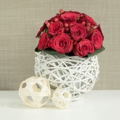 At the centre of the image, a lush dome of crimson red roses rises from a delicate white twig sphere, creating a wedding table centerpiece that feels both dramatic and perfectly composed, as though set for an evening celebration in a Tufnell Park venue. The roses are packed closely together, their velvety petals layered in tight spirals and soft curls, some still in bud and others just opening enough to hint at their inner depth. Nestled between the roses are clusters of deep red hypericum berries, smooth and glossy, like tiny beads of fruit that catch and reflect the light. A subtle rim of dark green leaves peeks out beneath the blooms, giving structure and a natural frame without stealing attention from the rich reds above. The flowers sit in a sculptural, spherical vase crafted from white-painted, interlaced twigs, the openwork pattern allowing glimpses of shadow and table beneath, lending a contemporary rustic feel that would look at home in a townhouse near Tufnell Park Road or a chic North London bistro. On the left side of the white tabletop stand two decorative balls woven from pale, natural-coloured wood, their gentle beige hue and crisscrossing strands mirroring the texture of the vase in a softer, more casual way. Behind, the background is softly blurred in light beige or taupe tones, perhaps a textile wall or subtle wallpaper, creating a calm, unobtrusive backdrop. The entire composition speaks of romance, thoughtful design, and quiet sophistication, ideal as a wedding reception centerpiece or romantic dinner arrangement arranged locally in Tufnell Park.