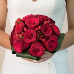 Resting lightly in the hands of a bride clad in white lace gathered close to Tufnell Park's leafy avenues, this radiant bouquet is a poetic expression of devotion. Dozens of deep red roses spiral together, their lush, velvety petals unfolding with both drama and gentle grace, tinted by the filtered afternoon light that so often bathes Tufnell Park's historic streets. Weaving between the blooms, vibrant red hypericum berries emerge like glistening gems, offering subtle textural relief and reflecting the joyful bustle of the local community. The tightly bound bouquet is shaped to perfection, ringed with glossy deep green foliage that forms a harmonious, sculpted collar-much like the carefully tended gardens found along Dartmouth Park Hill. The bride's polished nails and steady fingers cradle the stems, highlighting the bouquet's significance on her special day. Hints of floral fragrance and earthy undertones speak to the bouquet's freshness, recalling weekend strolls through Tufnell Park's artisan flower markets. In this heartwarming composition, the timeless allure of crimson roses and berries becomes a symbol of unity and abundance, a bold yet classic choice for those wishing to bring a touch of both romance and North London spirit to their wedding celebrations.
