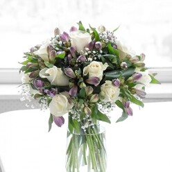 A sophisticated bouquet of creamy white roses, dusky lavender alstroemeria, and feathery white baby's breath is artfully arranged in a clear cylindrical glass vase, illuminated by soft daylight that suggests it rests by a window in a peaceful Tufnell Park living room. The white roses, fully opened yet still fresh, present a lush rosette of layered petals, their surfaces smooth and almost satin-like, lending the arrangement a sense of classic romance. Around them, alstroemeria blooms in shades of muted purple and lilac cascade outward, some petals just beginning to unfurl from their green sepals, others fully open to reveal fine streaks and veining, adding depth and visual intrigue. Sprays of gypsophila float among the larger flowers, like tiny white stars scattered through a pastel sky, softening the transitions and filling any gaps with gentle, airy texture. The surrounding greenery is deep and vibrant, with glossy leaves and svelte stems providing structure and contrast, as though freshly snipped from a garden bordering a quiet street near Tufnell Park station. Inside the clear vase, straight, cleanly trimmed stems are visible in the fresh water, showcasing the care with which the design has been built from the base up. The bouquet's overall profile is rounded and full, with each flower type thoughtfully placed so the colours flow seamlessly from cool whites to delicate purples. This arrangement gives an impression of calm optimism and heartfelt sincerity, ideal for sending a message of congratulations, offering solace during difficult times, or simply bringing an elegant, restful touch to a dining table or hallway console in a North London home.