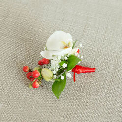 A single, artfully composed boutonniere rests on a pale oatmeal-coloured, woven fabric surface, the subtle weave of the material evoking the cosy interiors of Tufnell Park period homes near Tufnell Park station. At its centre, a flawless white Phalaenopsis orchid opens in a serene, sculptural form, each petal softly curved and slightly luminous against the neutral background. The orchid's throat glows with a delicate mix of creamy yellow and muted orange, drawing the eye to its intricate heart. Around this focal bloom, sprays of fine, white baby's breath scatter like tiny stars, giving the arrangement an airy, romantic lightness. Nestled close by, a vivid cluster of smooth, cherry-red berries brings an immediate sense of festivity, their rounded shapes and high shine suggesting holiday gatherings, winter weddings, and special evenings out along nearby Dartmouth Park Hill. A few pale green, berry-like buds offer a gentle contrast, hinting at growth and new chapters. Varied green leaves in rich, fresh tones weave through the flowers and berries, some broad and pointed, others slim and elongated, creating a natural frame that enhances the orchid's purity. At the base, the stems are carefully bound with a bright red satin ribbon, wrapped tightly and finished in a neat, compact knot with short, refined tails. The ribbon's sheen adds a celebratory accent, completing a piece that feels both modern and timeless. This small floral accessory seems perfectly suited for a groom, groomsman, or prom guest in Tufnell Park, a meaningful touch crafted with the same attention to detail you'd expect from a dedicated local florist.