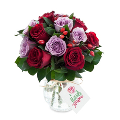 A lush, intimate bouquet composed of roughly equal parts deep crimson and burgundy roses paired with delicate lavender and lilac roses, each bloom opened to reveal layered, satiny petals that invite a gentle touch. The pairing creates a soft choreography of warm and cool tones: the intense reds bring passion while the cool mauves bring calm, together forming a balanced, romantic statement. Needle-like stems of dark green foliage - likely ruscus or salal - thread through the arrangement, giving structure and setting off the roses' colours with fresh contrast. Bright red-orange rose hips are dotted throughout, their glossy roundness and rustic texture offering lively punctuation against the velvet petals. The stems are presented in an antique-effect mercury glass vase, rounded at the base and tapering to a narrower neck; its silvered, crackled surface glints under diffused daylight, lending the bouquet an old-world elegance softened by the natural raffia tied around the neck. A small white rectangular tag is tied to the raffia, bearing a delicate floral sketch and space for a gift message - in practice this arrangement is often sent with a handwritten note for birthdays or heartfelt thank-you's. Prepared with local care for customers around Tufnell Park, Dartmouth Park and nearby Kentish Town, this composition - available for local delivery via Flowers Tufnell Park - reads as thoughtful, quietly luxurious, and perfectly suited to celebrations, anniversaries, or simply brightening a north London home.