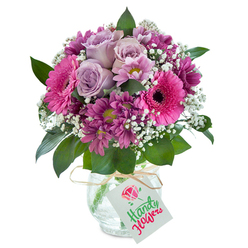 This radiant bouquet brings together the gentle romance of North London's floristry with the bustling life of Tufnell Park. The calm white background highlights vivid hues-a stunning interplay of lavender, fuchsia, and rich magenta flowers, artfully blended into a dense, rounded shape. At the heart, delicate pale purple roses exude understated elegance, softly opening petals inviting a breath of fresh fragrance reminiscent of early summer walks in Tufnell Park's quiet residential streets. Spirited, deep pink gerbera daisies radiate warmth and playfulness, their bold faces drawing the eye, much like the vibrant shopfronts on Fortess Road. Surrounding these are clusters of velvety plum chrysanthemums, their intricate layers and greenish-yellow centres providing both visual and tactile richness. Wisps of crisp white gypsophila interlace the arrangement, creating a cloud-like lift and sense of gentle movement-a floral translation of a breezy day in nearby Waterlow Park. Lush dark green foliage frames each bloom with a natural elegance. The bouquet rests in a clear glass vase marked by a distinctive crackled texture, tied simply at the neck with a strand of rustic raffia. From the raffia, a white rectangular gift tag dangles-ready to hold a personalised message-to complete the impression of a bespoke creation ready for delivery across Tufnell Park. Crafted with a professional eye for colour, scent, and texture, this design is ideal for celebrating milestones or sharing sincere affection within the local community.