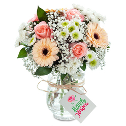 Visualise a gentle, restful morning in Tufnell Park, sunlight filtering across a bouquet that radiates both warmth and calm. Crystal-clear glass forms the vase, within which soft peach-pink roses unfurl their plush petals, an emblematic centrepiece. Peach-toned gerbera daisies bask nearby, their wide petals and textured brown centres lending the arrangement a sunlit brilliance reminiscent of neighbouring parklands like Whittington Park. Wisps of baby's breath meander between the main flowers, their airy white blooms reminiscent of pillows of cloud, while clusters of small chrysanthemums with verdant green centres inject sprightly freshness. A scattering of deep green foliage, perhaps eucalyptus, nestles among the pastels, grounding the flowers and offering subtle texture. The arrangement is sealed with a rustic raffia tied at the vase neck and a clean, rectangular white tag-a familiar mark in Tufnell Park's bustling independent flower shops. This bouquet embodies heartfelt affection, thoughtful celebration, or gentle support, mirroring the welcoming, community-centric spirit that runs through Tufnell Park's streets.