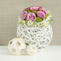 On a smooth white surface, a delicately crafted centerpiece seems to hover between rustic charm and modern design, capturing the kind of quiet elegance you might find at a relaxed Tufnell Park wedding reception or a stylish dinner party off Brecknock Road. The vase itself is a rounded, openwork sphere made from white, twig-like strips woven into a loose lattice, giving it the character of a contemporary wicker basket polished to a soft, chalky finish. Nestled inside this sculptural vessel is a lush dome of roses in gentle shades of blush pink and cool lavender, each bloom fully open and revealing layers of silky petals with subtly darker pink edges. Tucked among the roses, vivid green button chrysanthemums add a lively spark, their tight, spherical heads introducing both texture and a refreshing, garden-like energy. At the base of the arrangement, small white filler flowers cluster together, softening the outline of the bouquet and creating a feathery collar that hints at movement, as though the flowers have just been set down. To the left, two pale wooden or wicker spheres with an airy, crisscross pattern mirror the vase's woven theme, reinforcing a sense of craftsmanship and natural materials. The background is a gently blurred expanse of beige, like textured fabric or linen, providing warmth without distraction and allowing the colours of the flowers to stand out. The lighting is mild and even, picking up the delicate curls of each petal and the intricate twists of the wicker, evoking the skilled, attentive touch of a local florist accustomed to styling centerpieces for intimate gatherings around Tufnell Park, Kentish Town, and the surrounding neighbourhoods.