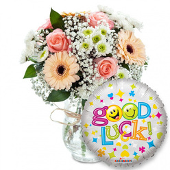 Here is a bright, encouraging arrangement that reads like a hand-delivered note of support: a lush floral bouquet set slightly left in a clear glass vase, tied simply with twine so the blooms feel both artisanal and approachable. The palette is gentle and hopeful - powder-pink roses with tightly coiled petals sit beside warm peach-to-orange Gerbera daisies with rich dark brown centres, their flat faces offering friendly focus. Soft clouds of white baby's breath (Gypsophila) spill through the composition like frothy lace, while small clusters of white and lime-green chrysanthemum-like florets bring a crisp, energetic counterpoint. Glossy deep-green foliage frames the flowers and adds shadow and structure under soft, even lighting. Dominating the foreground to the right, a round silver foil balloon shimmers with scattered colourful motifs - hearts, stars and tiny confetti dots in bright blue, pink, yellow and green - and proclaims 'Good Luck!' in playful, multicoloured lettering. The word 'Good' uses two yellow smiley faces for the o's, one with a wink and one with a cheeky tongue-out expression, which lends the ensemble a wink of mischief and warmth. A small KALEIDOSCOPE brand mark appears at the balloon's base. This composition feels perfect for sending encouragement to students finishing exams, someone starting work near Junction Road or a friend leaving for a new chapter; it reads as an uplifting, locally minded gift set that combines tender floral scent with buoyant visual optimism.