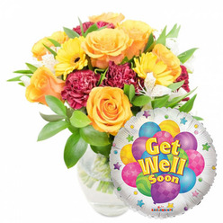 A comforting, cheerful get-well display arranged for bedside or doorstep delivery within Tufnell Park and surrounding streets. The bouquet sits slightly left of center in a clear glass vase, water and stems visible, crafted to feel both abundant and approachable. Central to the design are large, opening roses in warm orange and honey-yellow tones whose velvety petals suggest a soft citrus perfume; they form the visual heart of the arrangement. Sunny yellow gerbera daisies radiate optimism with bold, round faces, while ruffled maroon-to-burgundy carnations add tactile depth and a slower, textured rhythm among the brighter blossoms. Sprays of small white alstroemeria lift the composition with delicate marks of lightness; their fine petals introduce a gentle contrast against the stronger colours. A rich layer of glossy green foliage frames and supports the flowers, creating a sense of shelter and vitality. To the lower right, a metallic silver Mylar balloon subtly overlaps the bouquet, its reflective field featuring a cluster of illustrated balloons in vivid blues, purples, pinks, greens and yellows, some patterned with dots and stripes. The balloon's cheerful, shadowed yellow-and-blue lettering reads Get Well Soon, and small starburst motifs scatter around the border; a tiny KALEIDOSCOPE logo is visible at the rim. As arranged by Flowers Tufnell Park, this pairing of blooms and balloon is intended to brighten a recovery room or bring neighbourly warmth along Junction Road or Dartmouth Park walks.