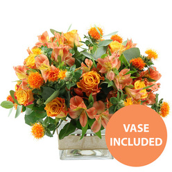 A rich, warm-toned bouquet is captured against a crisp white backdrop, its colours echoing the glow of an autumn evening over Tufnell Park. Dominating the arrangement are opulent orange roses, some with petals that gently shift to golden yellow at their centers, giving each bloom a softly illuminated look. Interwoven with them are sunny yellow garden roses, their densely layered petals forming plush rosettes that feel almost touchably soft. Between these showpieces, clusters of peach and light orange alstroemeria-Peruvian lilies with their slender, slightly flared petals-add a delicate rhythm, each blossom marked by fine streaks and speckles in deeper tones. Scattered throughout the bouquet, small, spiky, ball-shaped orange blooms, similar to safflower or thistle, create an intriguing texture, like tiny bursts of fireworks nestled among the more traditional flowers. The entire composition is cradled in a base of lush foliage: rounded, silvery eucalyptus fronds and darker green leaves that provide contrast and structure, enhancing the bouquet's sense of depth and movement. All the stems gather neatly into a clear, square glass vase, the cool transparency revealing the cleanly cut stems and water inside. Around the middle of the vase, a strip of natural beige burlap or jute is wrapped with care, adding a rustic, handcrafted touch that would suit a characterful home near Tufnell Park Fields or a stylish office close to Holloway Road. A bold orange circle appears at the bottom right of the image, with the words VASE INCLUDED in clear white capital letters, signalling that this generous, joy-filled arrangement arrives as a complete, ready-to-display floral gift for birthdays, thank you gestures, or cheerful same-day deliveries across the local area.