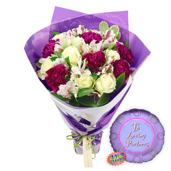 Set against a spotless white backdrop, this romantic bouquet and balloon pairing could easily be sitting in a Tufnell Park townhouse hallway, ready to surprise someone special. The flowers form a lush, rounded dome, with around a dozen creamy white roses at the centre. Each rose is tightly spiralled yet just beginning to open, their ivory petals smooth and softly luminous, suggesting a gentle, powdery scent. Interwoven with the roses are deep berry-toned carnations in shades of magenta and burgundy, their frilled petals densely packed, lending the arrangement a sense of richness and warmth. Between the larger blooms, white alstroemeria blossoms open gracefully, their long, narrow petals delicately marked with fine reddish-brown streaks and speckles, giving a subtle, almost secret detail for the eye to discover. Fresh green foliage, including some leaves edged in pale cream, billows around the flowers and peeks between stems, adding contrast and echoing the leafy calm of local streets around Tufnell Park station. The bouquet is wrapped with a florist's precision: an inner layer of clean white paper gives structure, while an outer skin of crinkled, translucent purple cellophane fans up around the blossoms, catching the light and bathing the edges of the petals in soft violet hues. At the base, a slim bright green satin ribbon is tied in a neat bow around the gathered stems, a small light brown tag dangling from the knot, ready for a personal note. To the right, resting on the same white surface, lies a round metallic purple balloon with a scalloped, rosette-like edge. Its lighter central panel carries the words "To Loving Partners" in flowing script, a tender message for couples celebrating anniversaries or shared milestones. A small jagged-edged rainbow holographic sticker adds a final playful sparkle, completing a gift that feels tailored for intimate celebrations across Tufnell Park.
