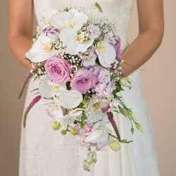 Captured in an elegant, natural-light setting near the heart of Tufnell Park, this captivating image features a bride holding a stunning cascading bridal bouquet-a floral composition radiating softness, movement, and local charm. The bridal gown, crafted from delicate white fabric adorned with intricate lace, provides a classic background for the show-stopping flowers she gently hugs to her torso. The bouquet's design is both contemporary and timeless, with pure white Phalaenopsis orchids forming a luminous, luxurious focal point, their petals glowing softly against mixed pastels. Around these, clusters of old-fashioned blush pink roses and ethereal lavender roses are placed at varied heights, their lush velvety petals conjuring the fragrance of summer mornings in nearby Waterlow Park. Wisps of snowy baby's breath create an airy, frothy layer, reflecting a sense of hopeful anticipation and gentle love. Here and there, smaller purple and lilac blossoms-possibly stock or locally grown sweet pea-bring further dimension and subtle aroma. Long amaranthus trails hang boldly, their magenta hues and feathery texture lending movement and contrast, while slender green leaves and unopened buds subtly nestle amongst the flowers. The overall palette of creamy whites, soft pinks, and twilight purples speaks to romantic strolls through Tufnell Park at dusk. Perfect for inspiration or local floral design, this bouquet's artistry reflects the neighbourhood's vibrant, creative spirit and a commitment to crafting unforgettable wedding moments.