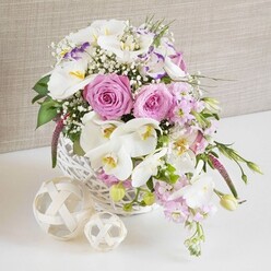 A beautifully composed floral centerpiece, ideal for a wedding or engagement celebration in Tufnell Park, sits proudly in a round white wicker basket placed on a crisp white table. The eye is drawn first to the lush pink roses, fully open and spiralling outwards, their soft blush tones radiating warmth and gentle romance. Cradling these roses, sculptural white Phalaenopsis orchids arc gracefully, their broad petals smooth and luminous against the basket, with golden-yellow throats that hint at exotic luxury. Interwoven between the larger blooms, frilled white Lisianthus flowers, some kissed with a faint purple edge, add depth and refined detail, while stems of pale pink stock spill lightly to the sides, bringing a sense of height and movement that feels almost like a breeze drifting through nearby Tufnell Park gardens. Fine sprays of white baby's breath are threaded delicately throughout, creating a haze of tiny blossoms that soften the transitions between flowers and lend the arrangement a dreamy, bridal quality. Glossy green leaves and a few tender buds emerge here and there, grounding the pastel palette with a touch of natural freshness. Beside the basket, two decorative balls woven from pale rattan or wicker rest on the same white surface, their interlaced strands mirroring the basket's texture and enhancing the rustic-yet-refined style. Behind it all, a softly textured background in pale taupe or beige keeps the scene calm and understated, allowing the intricate flowerwork to shine. The overall impression is one of thoughtful craftsmanship and quiet celebration, the sort of centerpiece that might anchor a wedding table in a local Tufnell Park restaurant or a thoughtfully styled home gathering.