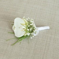 A refined white orchid boutonniere is captured lying on a pale beige, woven fabric that resembles softly textured linen, its subtle threads forming a calm, neutral backdrop like the interiors of a classic Tufnell Park apartment. The focal bloom is a flawless Phalaenopsis orchid, its petals smooth and gently curved, opening in serene layers of pure white that seem to glow against the understated background. At the centre of the orchid, a soft touch of buttery yellow nestles within the throat, lending the flower a gentle warmth and a sense of life and movement, as if it has just been carefully prepared in a local studio before a ceremony near Dartmouth Park. Surrounding the orchid, delicate sprays of baby's breath create a halo of tiny white florets, so light and airy they resemble a fine mist, cushioning the central bloom and adding a romantic, cloud-like texture. Here and there, wisps of slender green foliage peek out, their feathery lines and fresh, vivid colour suggesting the quiet greenery of nearby parks and gardens, bringing a natural balance to the design. The stems are meticulously aligned and bound with a strip of crisp white satin ribbon, wrapped smoothly and finished with a small, neat bow that rests close to the base of the flowers, indicating it is ready to be pinned to a jacket lapel. The satin's faint sheen softly echoes the orchid's natural gloss, tying the whole piece together. Crafted in the thoughtful, understated style often requested by couples marrying around North London, this boutonniere feels perfect for a groom, groomsman, or cherished guest, reflecting purity, commitment, and a timeless sense of occasion. It would suit intimate civil ceremonies as well as more traditional church weddings, whether held close to Tufnell Park or in nearby neighbourhoods.