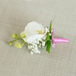 A single white orchid boutonniere rests gracefully on a pale oatmeal-toned linen surface, the woven texture beneath it providing a gentle, understated backdrop, like a freshly pressed suit laid out before an important day in Tufnell Park. The Phalaenopsis orchid at the centre is pristine and serene, with wide, gently curving petals that look almost porcelain-smooth, diffusing the soft light that falls evenly across the image. At its throat, a delicate blush of creamy yellow and a hint of pale pink add depth and warmth, drawing the eye inward to the flower's intricate structure. Surrounding the base of the orchid, fine sprays of white baby's breath spill outward in tiny, spherical blooms, creating a misty, almost cloudlike border that softens every edge. A few slender, bright green leaves extend from behind, their clean lines and glossy surfaces offering a refreshing contrast to the white flowers and neutral cloth, as if echoing the greenery found in nearby Tufnell Park and the gardens off Lady Margaret Road. On one side, two tightly closed lime-green orchid buds sit quietly on a slim stem, promising future blossoms and subtly suggesting growth and new chapters-perfect symbolism for a wedding, civil ceremony, or prom. The stems of the arrangement are gathered with care and wrapped in a narrow band of vivid fuchsia pink satin ribbon, finished in a neat, elegant bow whose satin sheen catches the light, lending a celebratory feel. The boutonniere exudes a calm, intimate mood, as though waiting on a dressing table before being pinned to a lapel for a special event close to home.