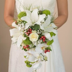 Cradled in the bride's hands, the cascading wedding bouquet becomes a centrepiece of natural grandeur, envisioned by skilled florists in Tufnell Park. Large white lilies take pride of place, their broad, star-shaped petals kissed with faint earthen freckles, creating a sense of serene luxury and classic charm. At the heart, a tightly coiled creamy white rose unfolds like a whispered promise, while trailing white Phalaenopsis orchids spill elegantly downward, capturing the movement of a gentle Islington breeze. Misty sprays of baby's breath encircle the main blooms, adding a soft, ethereal haze, while lively pops of lime-green chrysanthemums punctuate the design with cheerful, modern vibrance, echoing the renewal of nearby Tufnell Park gardens after rain. Scattered among the whites and greens, vivid clusters of red Hypericum berries gleam with energy-an accent that suggests joyful celebration, perhaps for a wedding reception on Fortess Road. Fine greenery, including slender, grass-like wisps and deeper foliage, frames the bouquet in layered texture and richness, giving the arrangement an organic, slightly wild grace. The bride's hands, visible just above the flowing ribbon, anchor the composition against the delicate lines of her ivory lace gown, the fabric's gentle patterns mirroring the bouquet's ornate artistry. Gentle daylight catches the satiny sheen of lilies and the velvety green of mums, revealing intricate detail at every glance. This bouquet radiates hope, fresh beginnings, and the artisan flair unique to Tufnell Park, perfectly suited for those seeking a balance between tradition and modern floral trends in North London ceremonies.