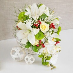 A luxurious wedding-style floral centerpiece rests on a white tabletop, its colours and textures echoing the calm, understated elegance often found in Tufnell Park homes and studios. The arrangement is cradled in a round white wicker basket, whose tightly woven strands create a natural, handcrafted base for the flowers above. Rising from it, several large white lilies unfurl their trumpet petals, showing softly freckled throats and delicate stamens dusted with pale pollen, evoking a subtle, sweet fragrance. At the heart of the design, a single cream-yellow rose glows softly, its petals forming a precise rosette that draws the gaze and adds warmth to the crisp whites. From the right-hand side, a cascade of white phalaenopsis orchids sweeps down in a graceful arc, each bloom smooth and porcelain-like, lending the bouquet a classic cascading or teardrop feel that would sit beautifully on a wedding top table in a local Tufnell Park restaurant or hall. The composition is enriched with bright green pom-pom blooms, likely small chrysanthemums, their rounded, textured heads giving a mossy softness amid the sleeker petals. Wispy grasses and feathery foliage reach outward, adding lightness and motion, while clusters of glossy red berries bring unexpected splashes of crimson, like tiny jewels buried among leaves. Sprays of white baby's breath weave through the design, filling the gaps with fine, cloud-like blossoms that enhance the airy quality. To the left of the main bouquet, two white woven decorative spheres sit quietly on the table, their open lattice pattern mirroring the basket and completing the scene. The soft beige backdrop and gentle lighting create an intimate, serene atmosphere, perfect for marking moments of love, joy, and togetherness across the Tufnell Park area.