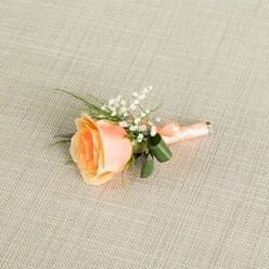 Displayed lengthways on a softly textured, light beige linen-style fabric, this peach rose boutonniere appears almost to glow with its own gentle warmth, like the sky just before sunset over the terraces of Tufnell Park. At the centre, a single rose is caught mid-bloom, its petals curling outward in a spiral of colour that moves from a warm, apricot-peach heart to creamy, pastel edges. Nestled against its upper curve, clusters of white gypsophila-baby's breath-form a frothy halo of tiny blossoms, each one a pinpoint of brightness against the rose's richer tones. Slim green blades of foliage and a pair of darker, broader leaves peek from behind, adding depth, movement, and a natural, garden-fresh feeling, as if the flowers had just been picked from a careful cutting patch behind a local townhouse off Dartmouth Park Hill. The stems are carefully gathered and wrapped in a smooth peach satin ribbon, its sheen catching the soft, even light that gently washes over the scene. The ribbon winds neatly down the stems, finishing in a small, unobtrusive knot that keeps the design compact and secure, ready to be pinned to a groom's or guest's lapel. The background cloth, with its fine woven grain, stays resolutely understated, letting the intricate form of the rose, the airy baby's breath, and the glossy greens remain the quiet stars. Crafted with the precision you'd expect from an experienced florist serving weddings and formal events around Tufnell Park, this boutonniere conveys a mood of intimate celebration and timeless romance.