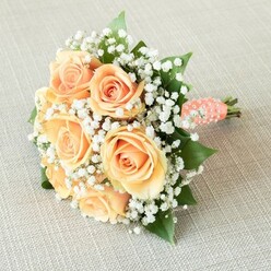 Resting quietly atop a finely textured, pale linen reminiscent of artisan market stalls along Tufnell Park Road, this exquisite bouquet reveals itself through soft sophistication and masterful design. Delicate peach and apricot roses-each bloom fuller than the next-anchor the arrangement, their petals gently unfurling amidst the gentle touch of daylight filtering through nearby London Plane trees. Surrounding the roses, feather-light sprays of baby's breath evoke the misty mornings of North London, adding a whisper of lightness and grace that lets the warmth of the central blossoms shine. Interspersed are defined, emerald-hued leaves that bring structure and life, finishing the arrangement with a sense of lush greenery typical of the area's hidden gardens. The stems are thoughtfully bound with a peach satin ribbon, its gentle sheen traced by a spiral array of tiny white pearls that offer a tactile hint of luxury-a nod, perhaps, to heirloom bridal styles beloved in Tufnell Park ceremonies. The bundle feels both substantial and delicately poised, intended for a bride or bridesmaid strolling past the charming cafés of nearby Fortess Road, or for anyone celebrating a moment marked by anticipation and love. The bouquet's fresh scent-tinged with dewy greenery and the promise of a new day-blends with memories, poised to remain long after petals have faded; a living tribute to tender celebrations in the heart of the community.