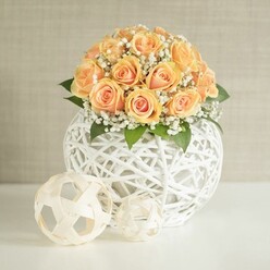In this image, a beautifully composed dome of around two dozen peach and apricot roses rises from a sculptural white sphere, creating a centerpiece that feels both luxurious and welcoming. Each rose is at a slightly different stage of bloom, some still tightly furled, others softly opening to reveal deeper, sunlit orange centres that transition to lighter, velvety edges. Fine sprays of white gypsophila thread through the roses, like tiny stars scattered in a pastel sky, lending a gentle, cloud-like softness that softens the compact shape. A touch of dark green foliage nestles low in the arrangement, giving a quiet contrast that keeps the colour palette grounded and natural. The flowers sit in a round, openwork vase crafted from white woven strips, almost like a contemporary wicker ball, its criss-crossing lattice adding a tactile, design-led element to the scene. Set on a pristine white surface, the vase is joined by two smaller decorative spheres, one slightly larger than the other, made from pale, natural-looking bands that curve and intersect into airy orbs. They echo the main container's texture and bring a sense of rhythm and balance. The backdrop is a soft, neutral beige wall with a subtle vertical grain, evoking the calm interiors of a renovated flat off Fortess Road in Tufnell Park. Gentle, even daylight falls across the arrangement, enhancing the warmth of the roses and the crispness of the white elements without harsh shadows. The overall mood is poised yet intimate, perfectly suited to an anniversary dinner at home, a wedding table by Tufnell Park station, or a refined gift arrangement prepared by a local florist who understands how to blend modern design with timeless romance.