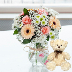 On a bright, clean white surface, a lovingly crafted floral arrangement and a small teddy bear are posed together in a way that feels like a private anniversary moment in a Tufnell Park living room. The bouquet is gathered in a clear glass jar vase, its simple shape allowing the flowers to take centre stage. Soft pink roses, just opening, form the romantic heart of the design, surrounded by peach-toned gerbera daisies whose velvety petals radiate from rich, dark brown centres. These sit within a generous collar of fresh white chrysanthemums and smaller daisy-like blooms, some with hints of lime green at their cores, adding a crisp, modern freshness. Wisps of baby's breath thread through the bouquet like a fine mist, giving it an airy, light quality, while a few discreet green leaves provide natural contrast without overpowering the gentle pastel palette. Around the neck of the vase, a piece of natural twine is tied casually yet thoughtfully, holding a little white tag that bears a pink heart illustration and the word Handmade in elegant green lettering, suggesting a personalised gift prepared with care by a local florist who knows the neighbourhood well. To the right, a cream-coloured teddy bear with fluffy, textured fur leans slightly towards the flowers. Its dark bead eyes, small brown nose, and neatly tied chocolate ribbon bow give it a sweet, nostalgic charm. Soft daylight, reminiscent of morning light drifting over Tufnell Park Road or nearby green spaces, washes over the scene, sharpening the colours while maintaining a feeling of calm. Together, bouquet and bear create a visual message of affection and reassurance, perfectly in tune with an anniversary celebrated quietly at home.