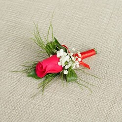 A meticulously crafted boutonnière rests on a pale, oatmeal-toned linen background, the neutral fabric providing a calm, tactile stage for a single, striking rosebud in shades of deep crimson-fuchsia. The rose itself is still in bud, its petals tightly coiled and satin-smooth, with a subtle gradient of colour that intensifies toward the centre, as though blushing with anticipation. Around this central bloom, airy tufts of white baby's breath are tucked in with care, their tiny blossoms resembling scattered snowflakes that form a gentle, cloud-like frame. Fine, grass-like fronds of bright green foliage arc gracefully behind and around the flowers, giving the piece lift and dimension, similar to the soft greenery that lines the pathways around Tufnell Park and Dartmouth Park on a fresh spring morning. At the base, all stems are cleanly aligned and firmly wrapped in a rich coral-red satin ribbon, the fabric coiled in neat, overlapping bands that catch the light with a subtle sheen, suggesting the polished elegance of formal attire. The ribbon is finished with a discreet loop or tiny bow, adding a tailor-made feel that hints at an artisan's patient hands rather than mass production. The entire boutonnière is angled slightly upward across the fabric, as if ready to be pinned to a lapel before a wedding ceremony at a local Islington venue or a special celebration near Fortess Road. Every element-the vivid rose, the crisp gypsophila, the feathery greenery, and the refined ribbon-works together to convey tenderness, celebration, and the quiet significance of the moments when a small flower at the heart of Tufnell Park becomes part of a much bigger memory.