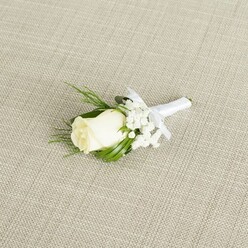 A refined wedding boutonniere rests at a diagonal angle on a pale beige background that resembles finely woven linen, the subtle texture of the fabric forming a neutral canvas for the florals. At its centre, a single creamy white rosebud stands proud yet understated, its smooth petals tightly layered and only just beginning to unfurl, giving the impression of a bloom at its freshest point. The rose is framed by delicate tufts of white baby's breath, each tiny, airy flower creating a cloud-like halo that softens the silhouette and adds a sense of lightness. Around these, sprigs of lush greenery stretch gracefully outward: some leaves are slim, pointed, and deep green, while others are more feathery, offering a gentle contrast of shapes and foliage textures. The stems of the rose, filler blooms, and greenery are carefully aligned and wrapped in a band of pure white satin ribbon, which has a gentle sheen that catches the soft light. Just below the main flower head, the ribbon is tied into a small, neatly formed bow, its tidy loops and short tails suggesting the confident hand of an experienced florist, perhaps one preparing boutonniere sets for a ceremony at a town house off Tufnell Park Road. The overall effect is one of quiet elegance and tradition: the white rose symbolises purity and new beginnings, while the baby's breath adds a whisper of romance. The diffused lighting ensures that every petal edge, leaf vein, and fold of ribbon is visible but never harsh, making the boutonniere appear timeless, ready to be pinned to a groom's lapel for a North London wedding or formal event steeped in understated style.